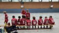 Kinder in roten Team-Shirts sitzen auf einer Bank in der Sporthalle, zu sehen sind die Rücken.