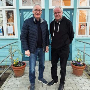 Foto, zwei Personen vor dem blauen Haus, sehen zur Kamera.